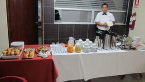 Una mesa de buffet con diversos alimentos y bebidas, un extintor en la pared, y una persona con camisa blanca y pajarita.
