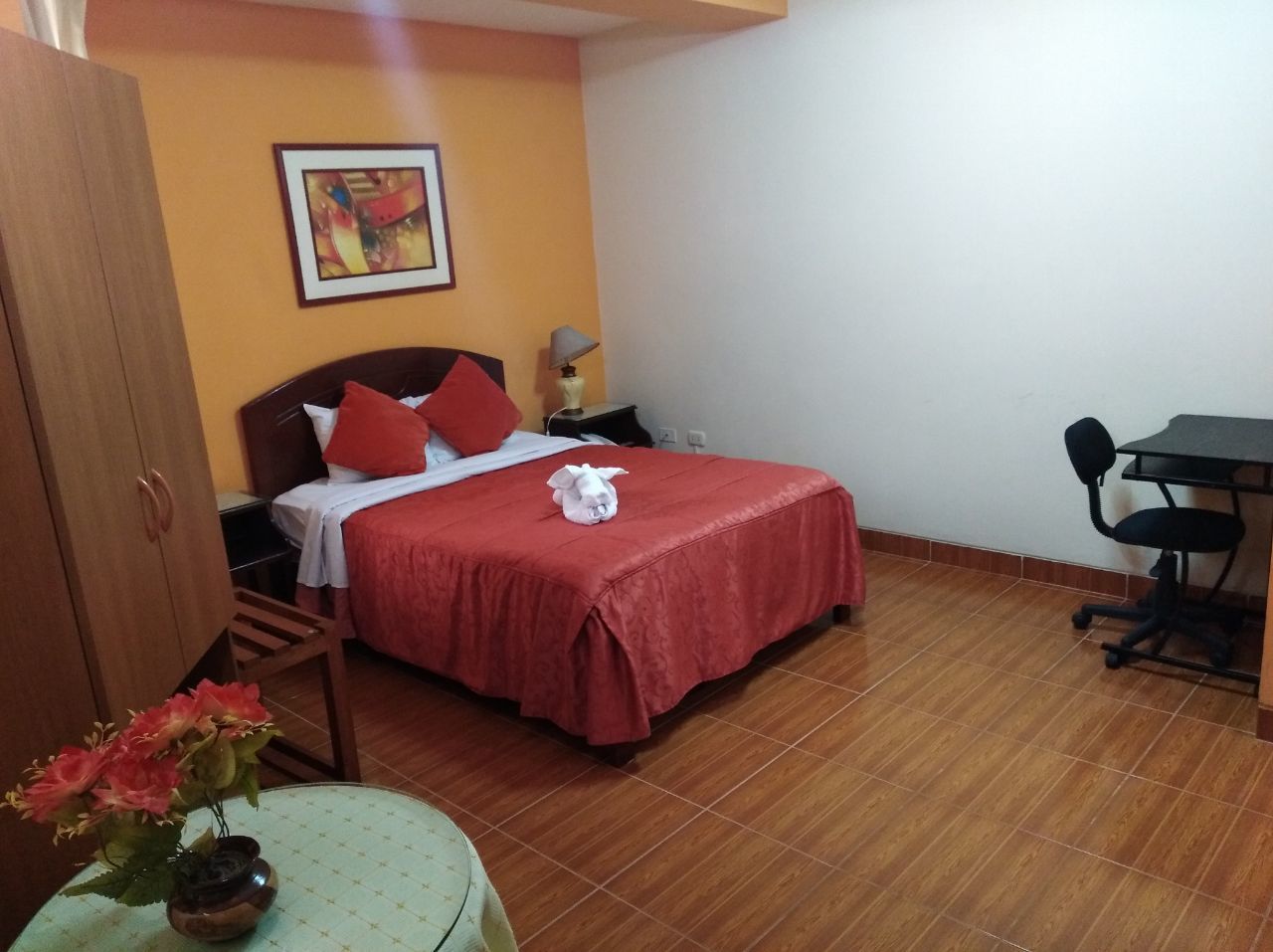Quarto com cama, escrivaninha, cadeira, guarda-roupa e um vaso com flores.