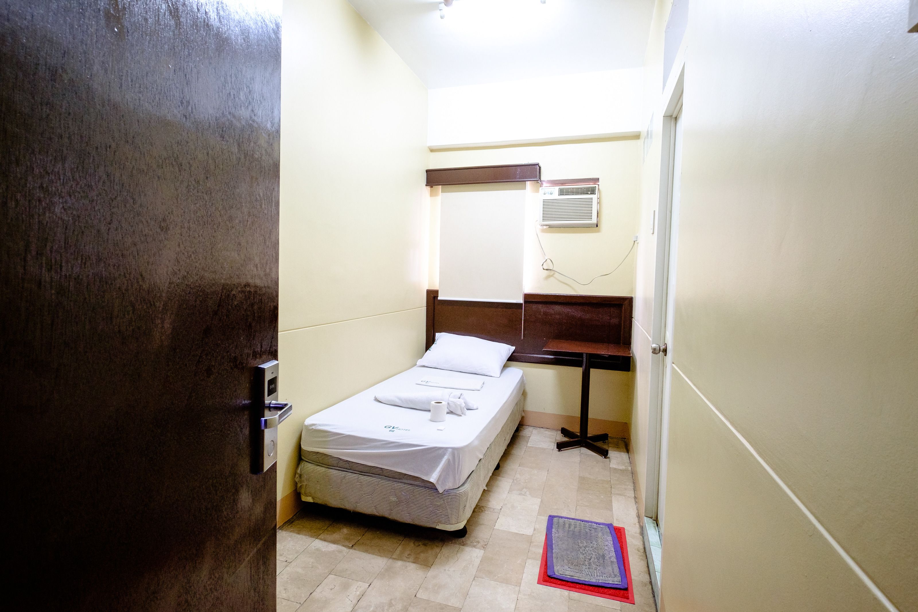A small, narrow hotel room with a single bed, a bedside table, a mirror, and a window with blinds.