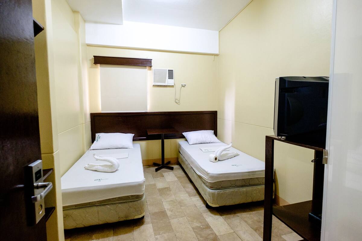 Une petite chambre d’hôtel avec deux lits simples, une tête de lit en bois, une petite table, un téléviseur et un climatiseur fixé au mur.