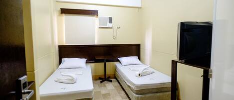 A small hotel room with two single beds, a wooden headboard, a small table, a television, and a wall-mounted air conditioning unit.