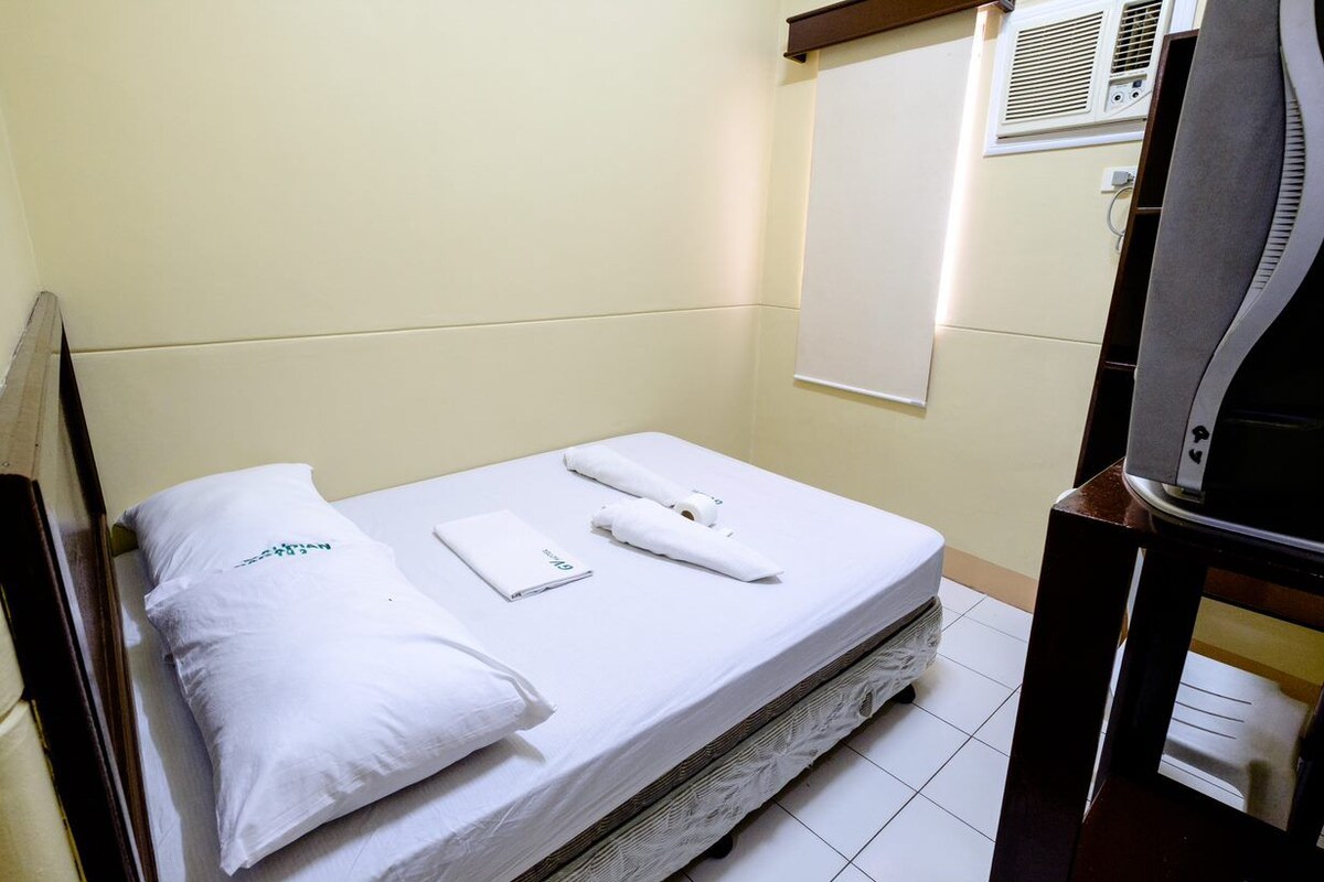 Cama de casal com roupa de cama branca, uma toalha dobrada e almofada em quarto com parede lisa e janela com persianas.