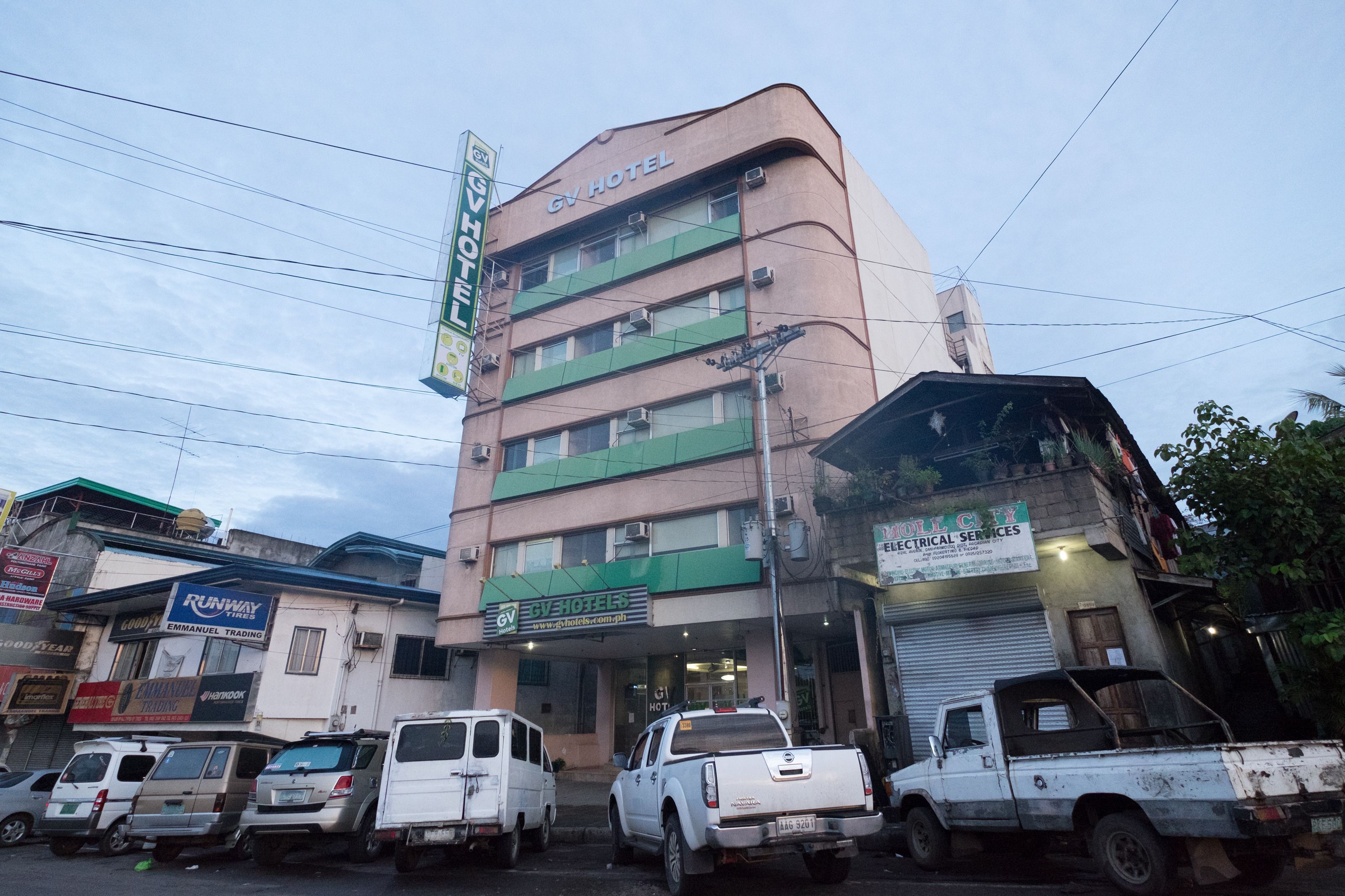 Un edificio de varios pisos con un letrero que indica GV Hotel. Varias unidades estacionadas frente al edificio, incluyendo una camioneta blanca.