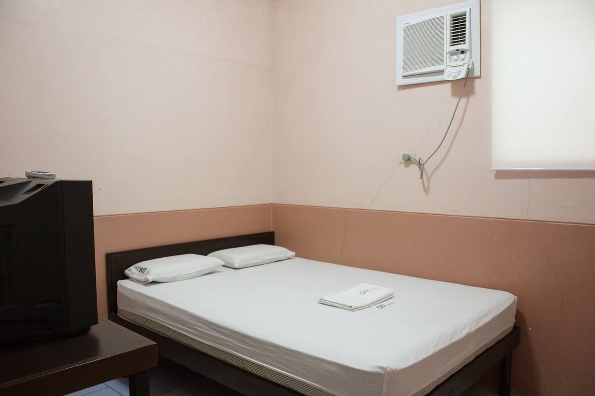 A small, single-bed room with a wooden headboard, a bedside table, a television, and a wall-mounted air conditioning unit.
