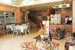 Un vestíbulo de hotel con mostrador de recepción, un área de comedor con mesas y sillas, y una escalera al fondo.