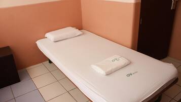 Un lit simple avec un matelas blanc et une serviette blanche pliée, le tout surmonté d’une tête de lit portant l’inscription « GV HOTEL ».