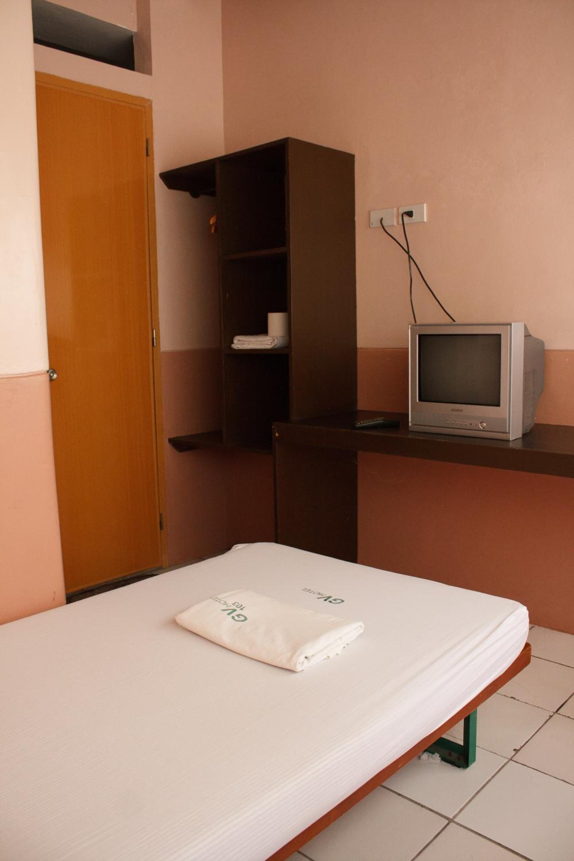 Una habitación pequeña con una cama individual, un armario de madera, un estante con televisor y un enchufe en la pared.