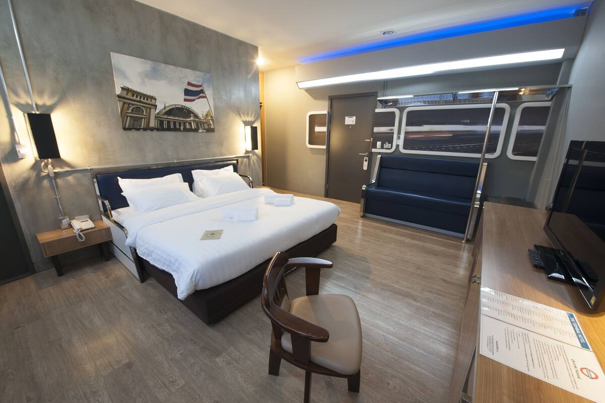 A hotel room with a bed, bedside table, chair, desk with computer, and a view of a train.