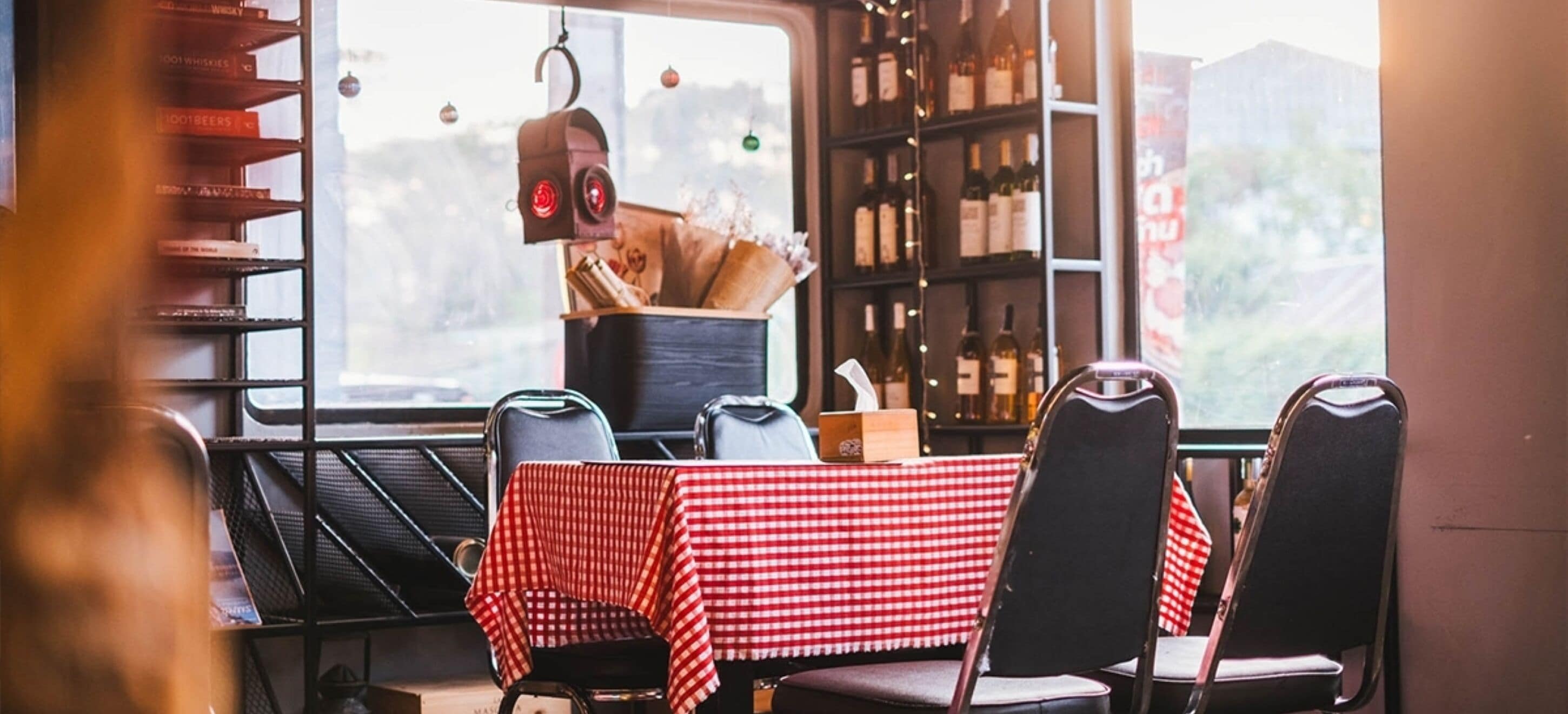 A restaurant interior with a red and white checkered tablecloth, black chairs, and a bookshelf filled with books and wine bottles.