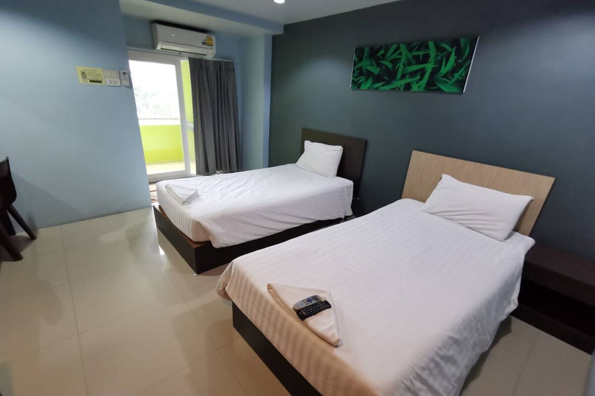 A hotel room with two beds, a large window with curtains, an air conditioning unit, and a green abstract painting on the wall.