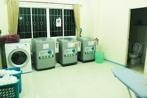 A laundry room with a front-loading washer, two LG TurboSpin dryers, a blue laundry basket, and a blue ironing board. - Able Resident Hat Yai (Hat Yai)