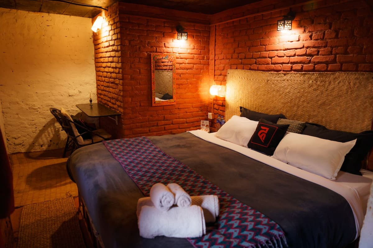A room with a brick wall, a bed with white and black bedding, a small table with a chair, and a wall-mounted light fixture.