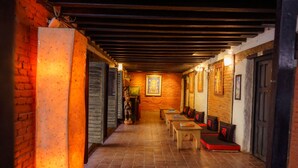 A narrow hallway with brick walls, wooden beams, and tiled flooring. There are benches and tables along the walls, and a statue is visible in the center. - Hira Guest House (Lalitpur)