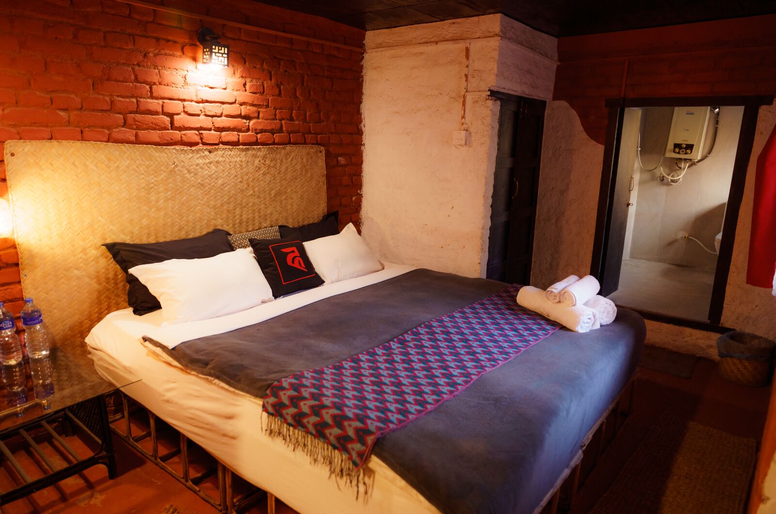 Een hotelkamer met een bakstenen muur, een bed met een geborduurde deken, kussens en een handdoek op het bed. Er is een raam met uitzicht op een douchegedeelte.