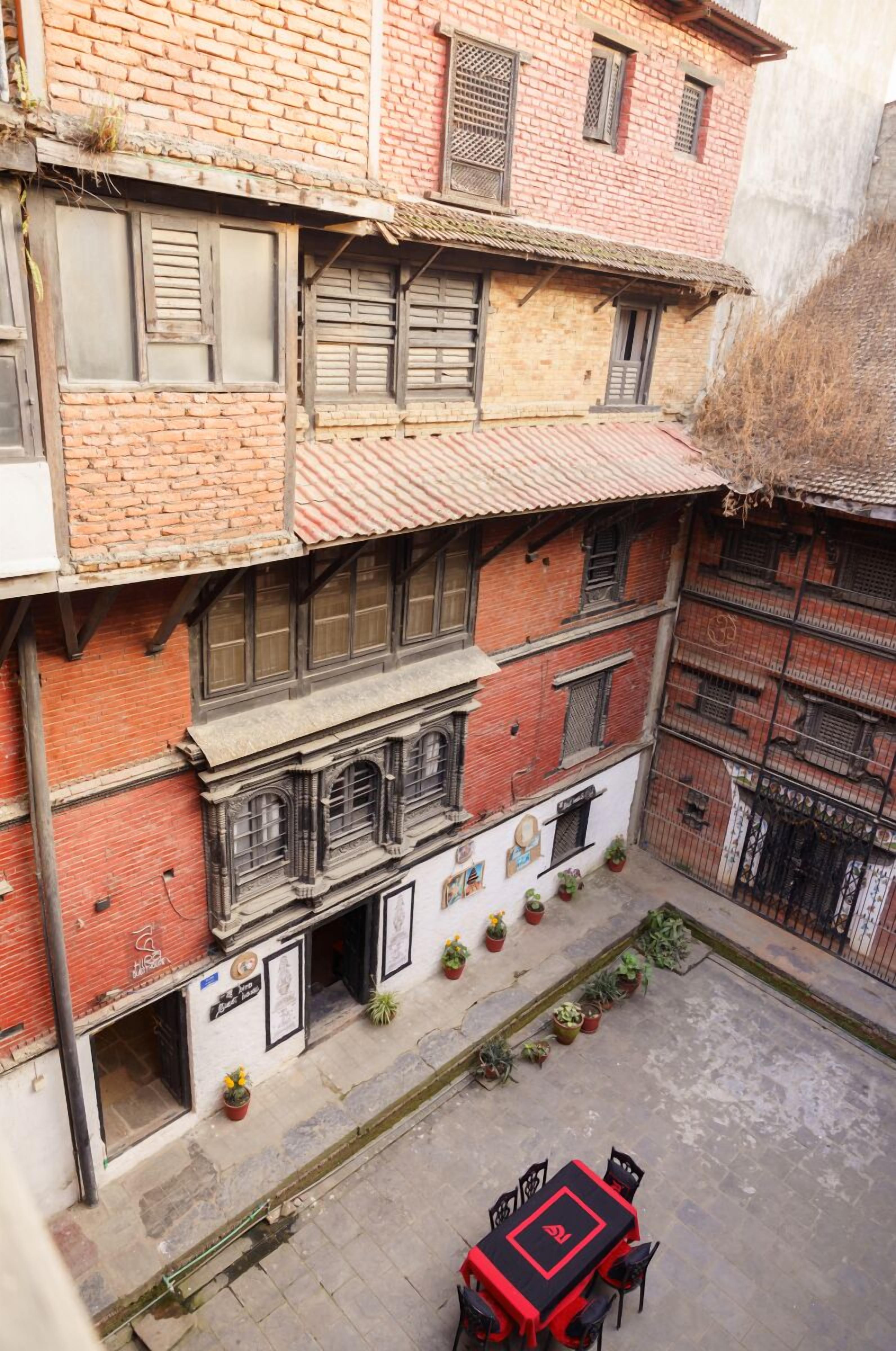 Un edificio de varios pisos con un patio interior. El edificio tiene múltiples ventanas con contraventanas, algunas de las cuales están abiertas. Hay un pequeño espacio al aire libre con una mesa y sillas. El patio tiene plantas en macetas y un camino pavimentado.