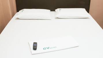 Uma cama de solteiro com colchão branco e cabeceira preta. Um travesseiro branco e uma toalha branca dobrada estão sobre a cama. Um controle remoto e um cartão branco com a inscrição 'GV HOTEL' também se encontram na cama.