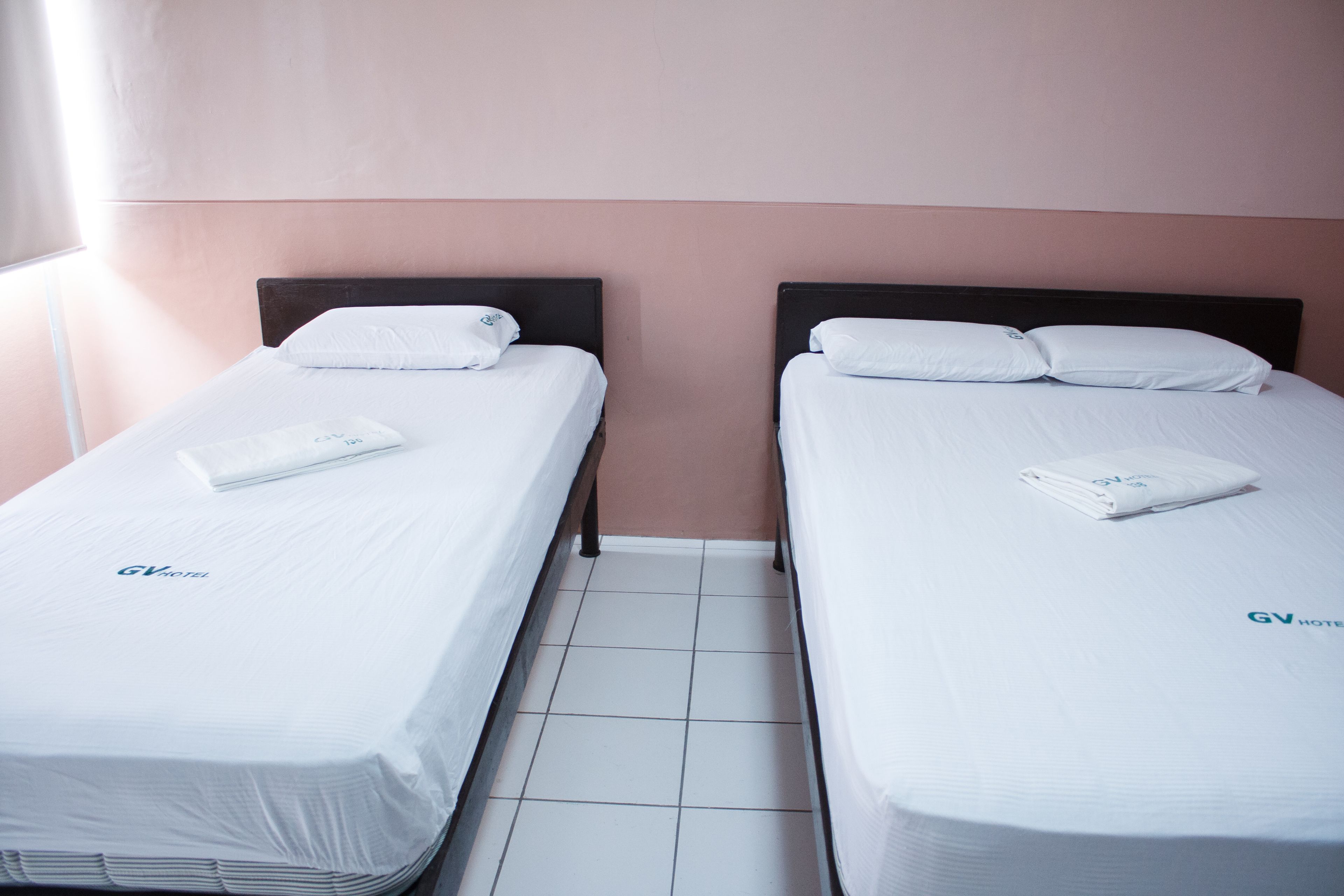 Deux lits simples avec du linge de lit et des oreillers blancs, un sol carrelé et un mur sobre avec une fenêtre sur le côté gauche.