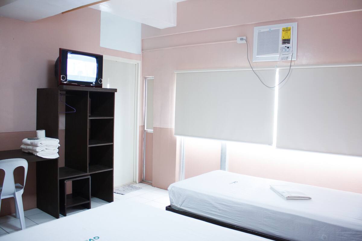 Ein Schlafzimmer mit Bett, Schreibtisch mit Stuhl, Fernseher auf einem Regal und einem Fenster.