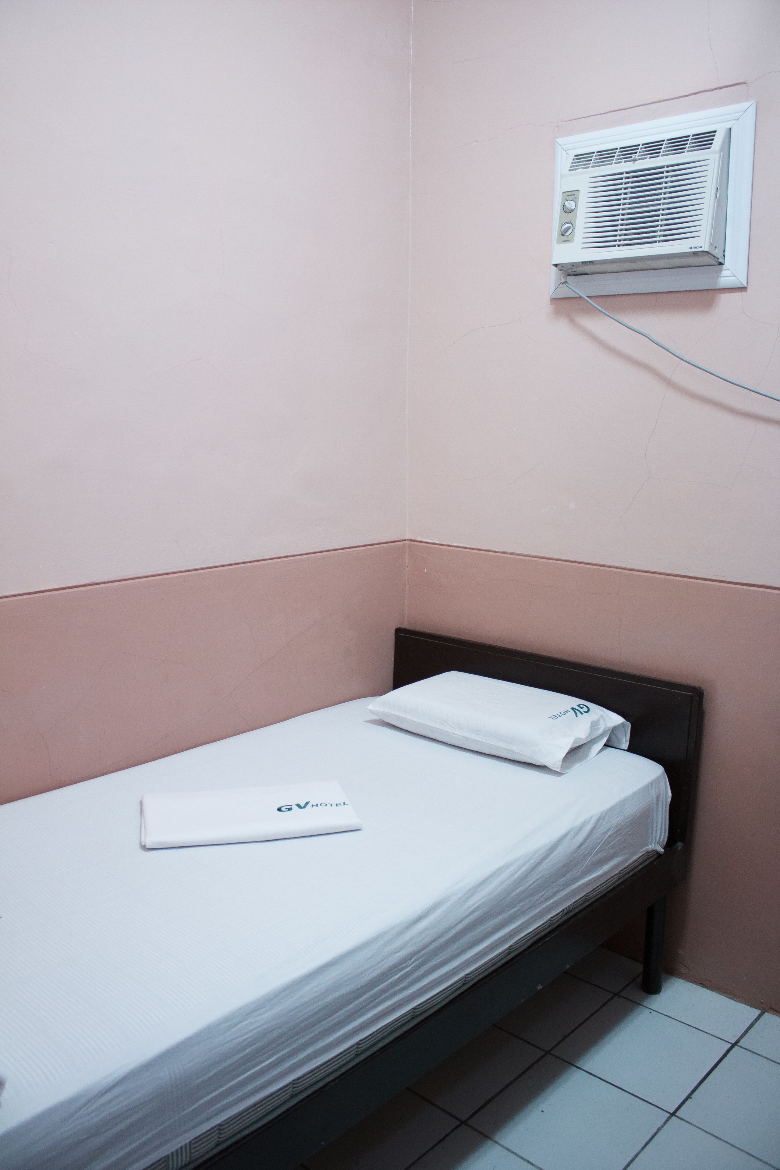 Une petite chambre avec un lit simple, une couette blanche, un oreiller blanc et une serviette blanche. Un climatiseur mural est fixé au-dessus du lit.