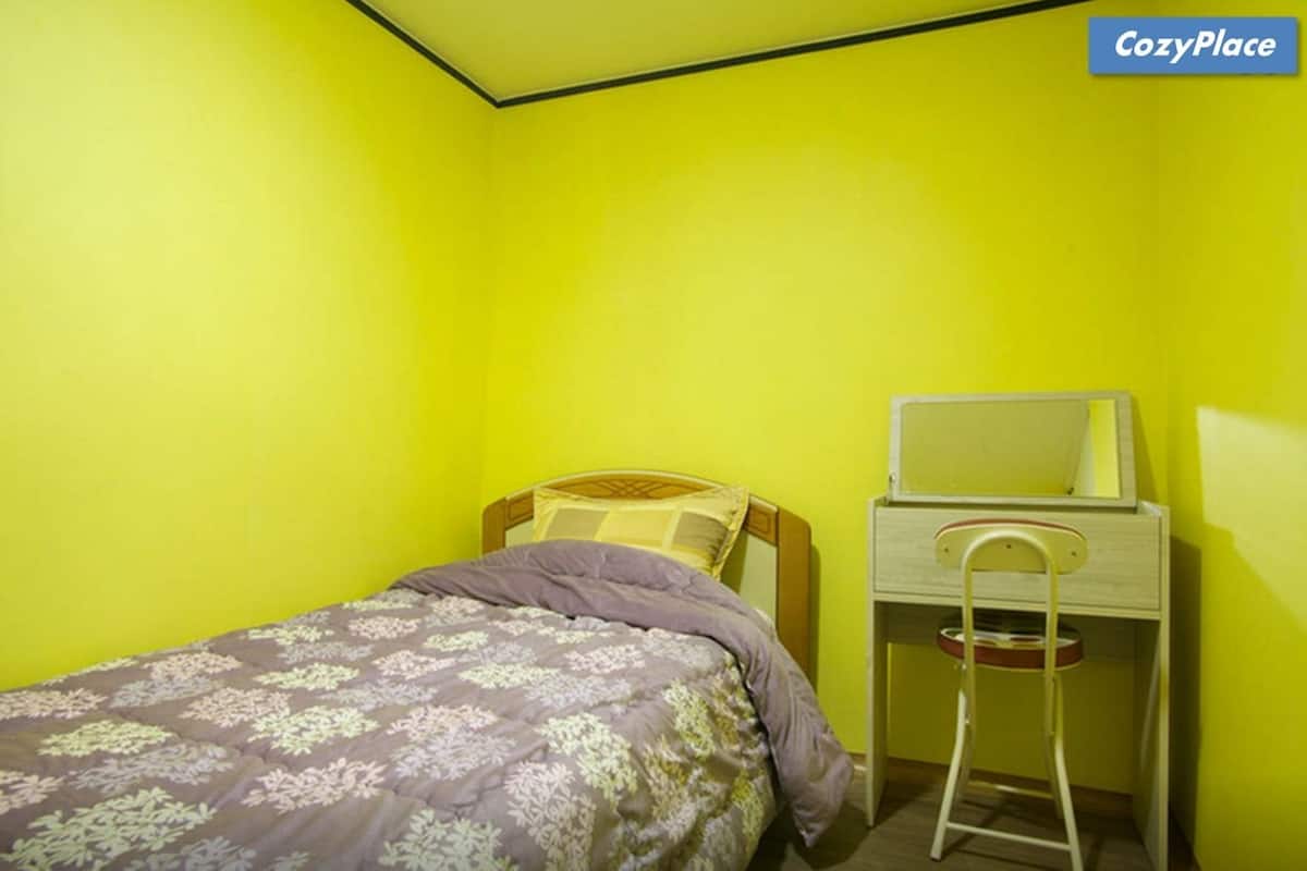 A small, brightly colored room with a single bed, a nightstand, and a chair.