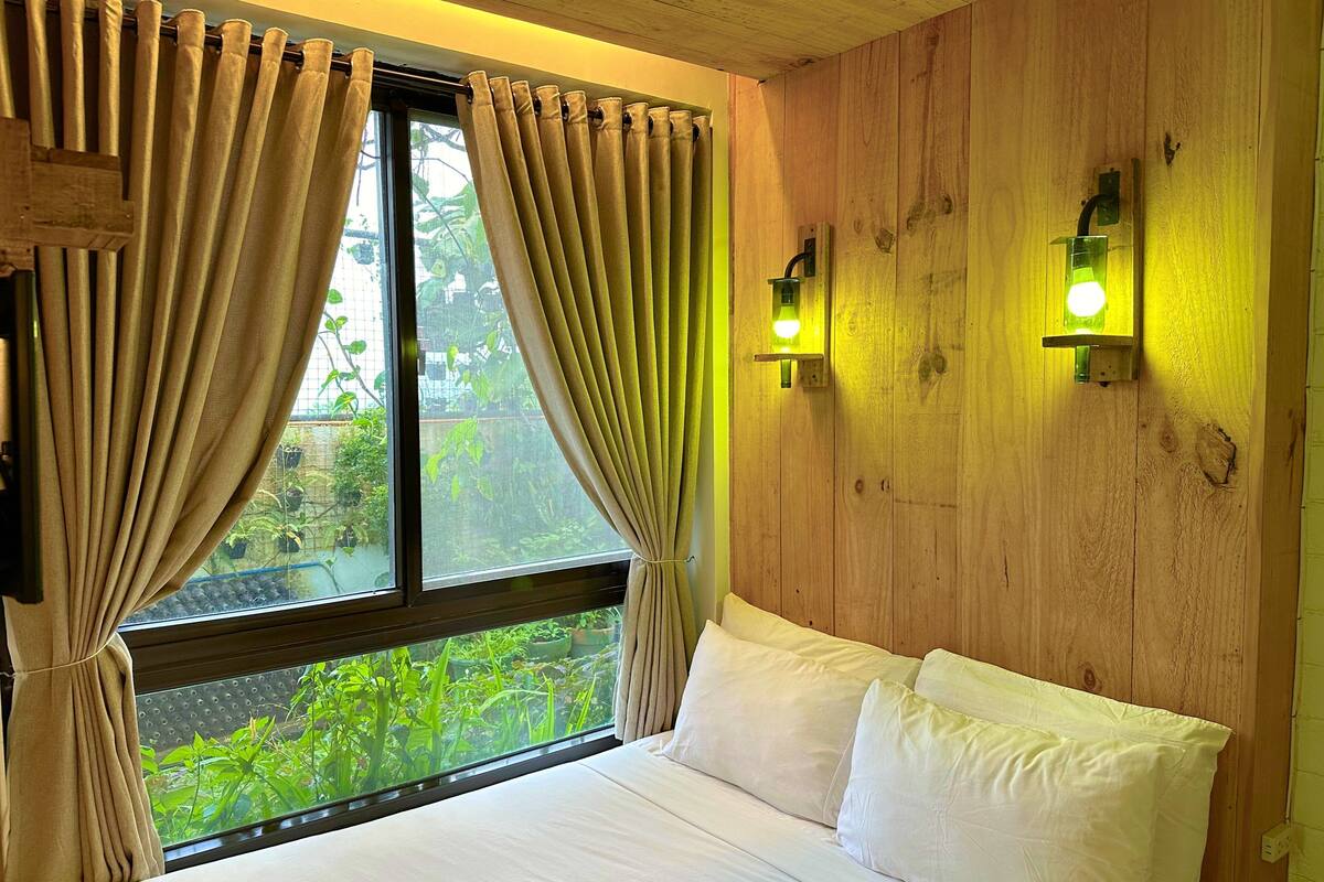 Een slaapkamer met een houten wand, een bed met wit bedlinnen en gordijnen. Aan weerszijden van het raam bevinden zich twee wandlampen.