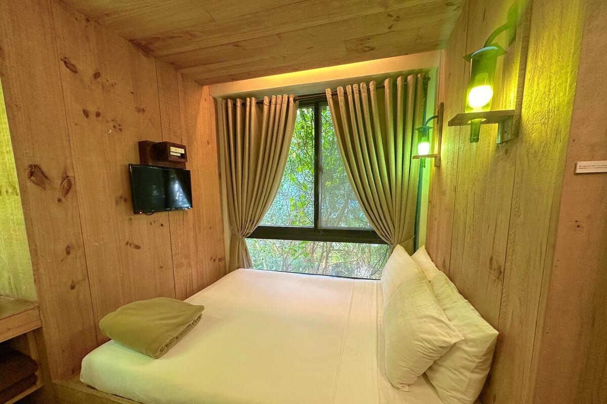 Une pièce en bois avec un lit, des rideaux, une lampe fixée au mur et une fenêtre donnant sur des arbres.