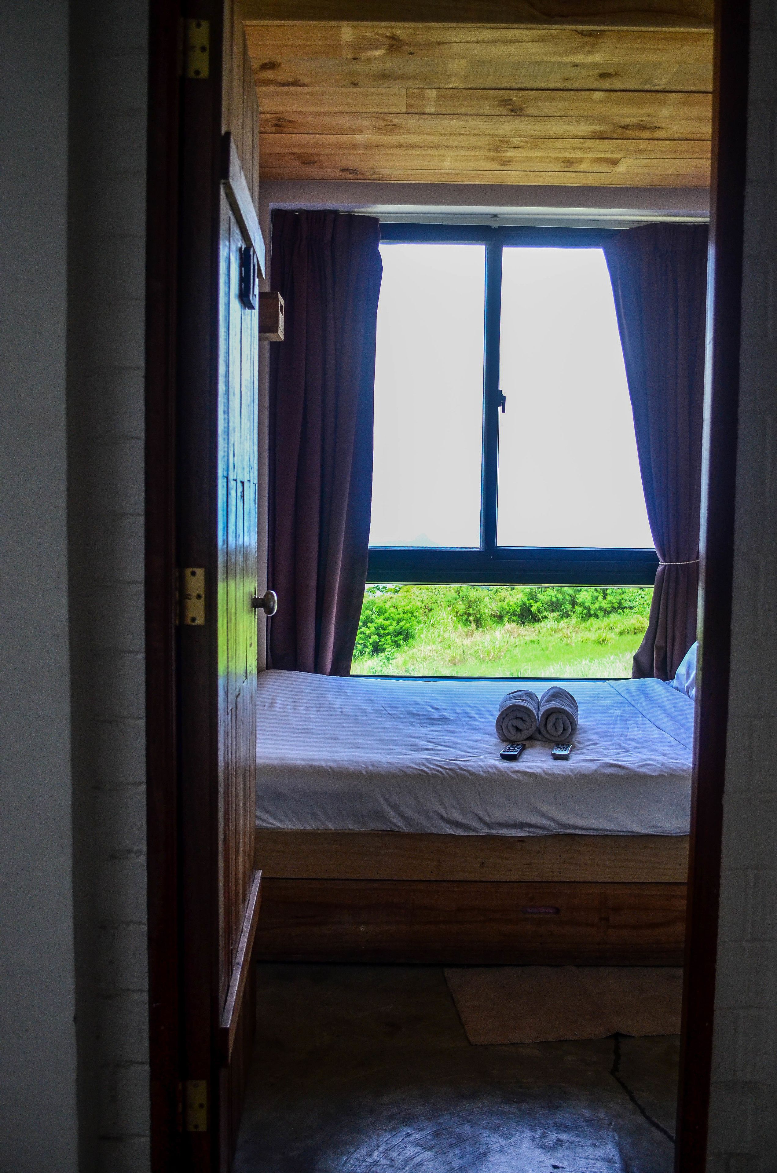 A bedroom with a wooden ceiling, a large window with curtains, a bed with a rolled-up towel and remote control, and a view of greenery outside.
