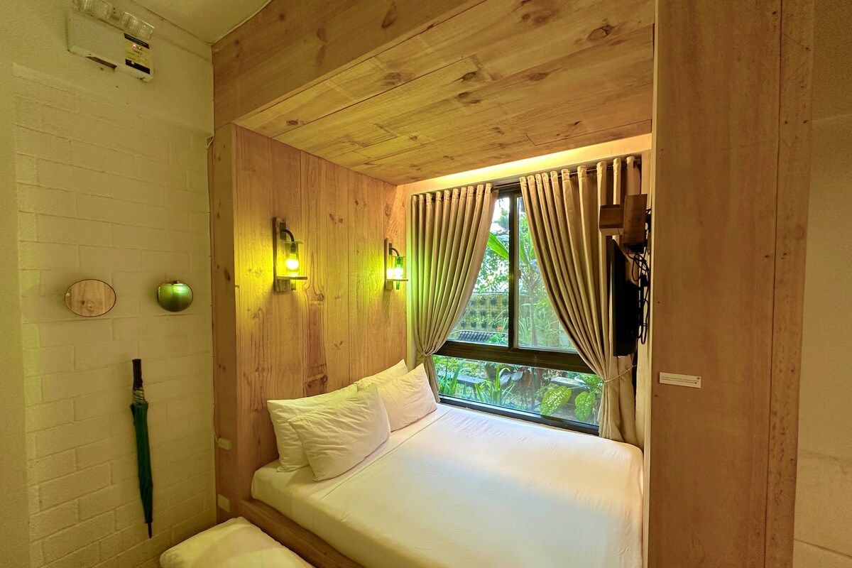 Een kamer met houten lambrisering, een bed, een raam met gordijnen, een wandlamp en een groene paraplu aan de muur.