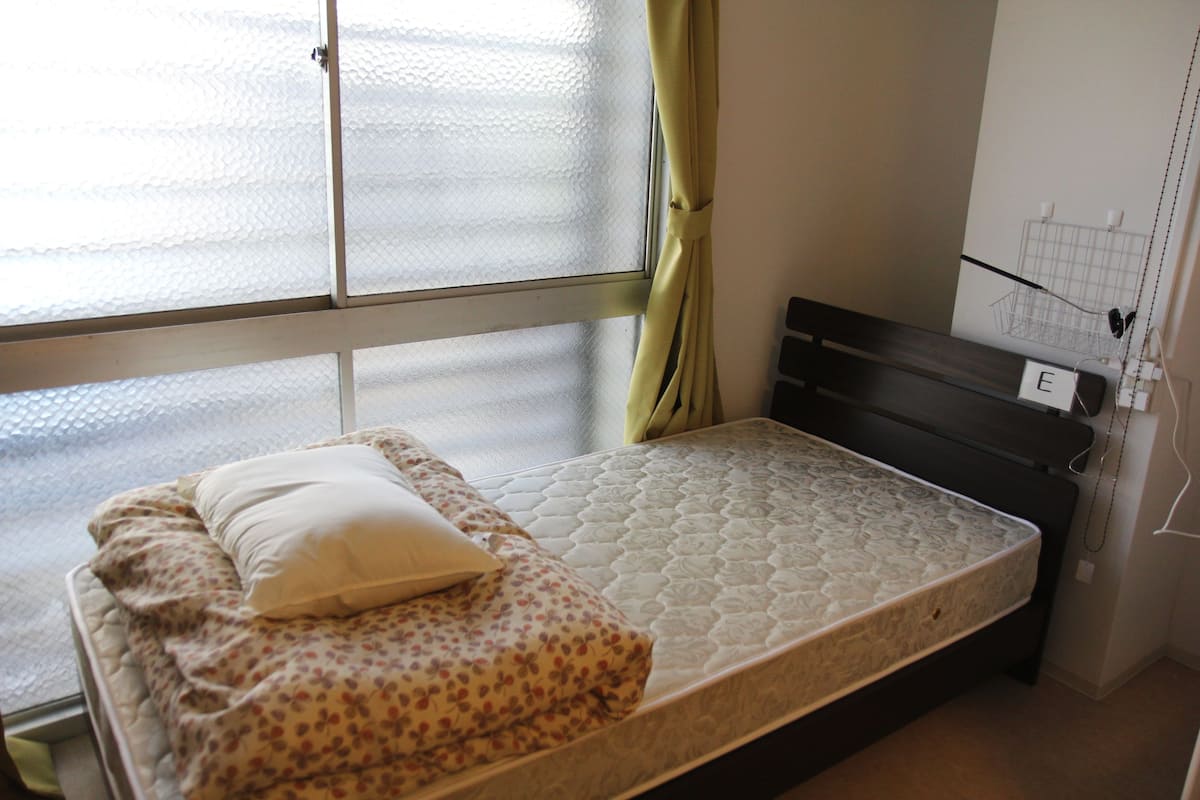 A small, single-bed room with a mattress, pillows, and a window with a curtain.