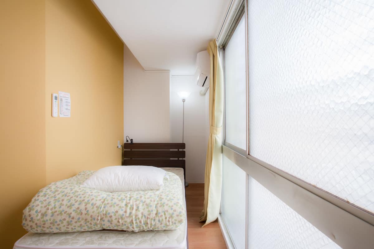 A small, single-bed room with a wooden headboard, a bedside table, and a window with curtains.