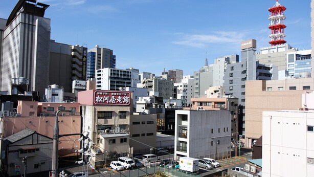 Un paisaje urbano con varios edificios, incluyendo un hotel con un letrero en un idioma extranjero, y una torre roja al fondo.