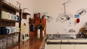 Cuina amb una estant de fusta que conté tasses, un brossa i una olla. Hi ha una calcomania mural d'un gat i sushi, i un rentapeus amb plats.