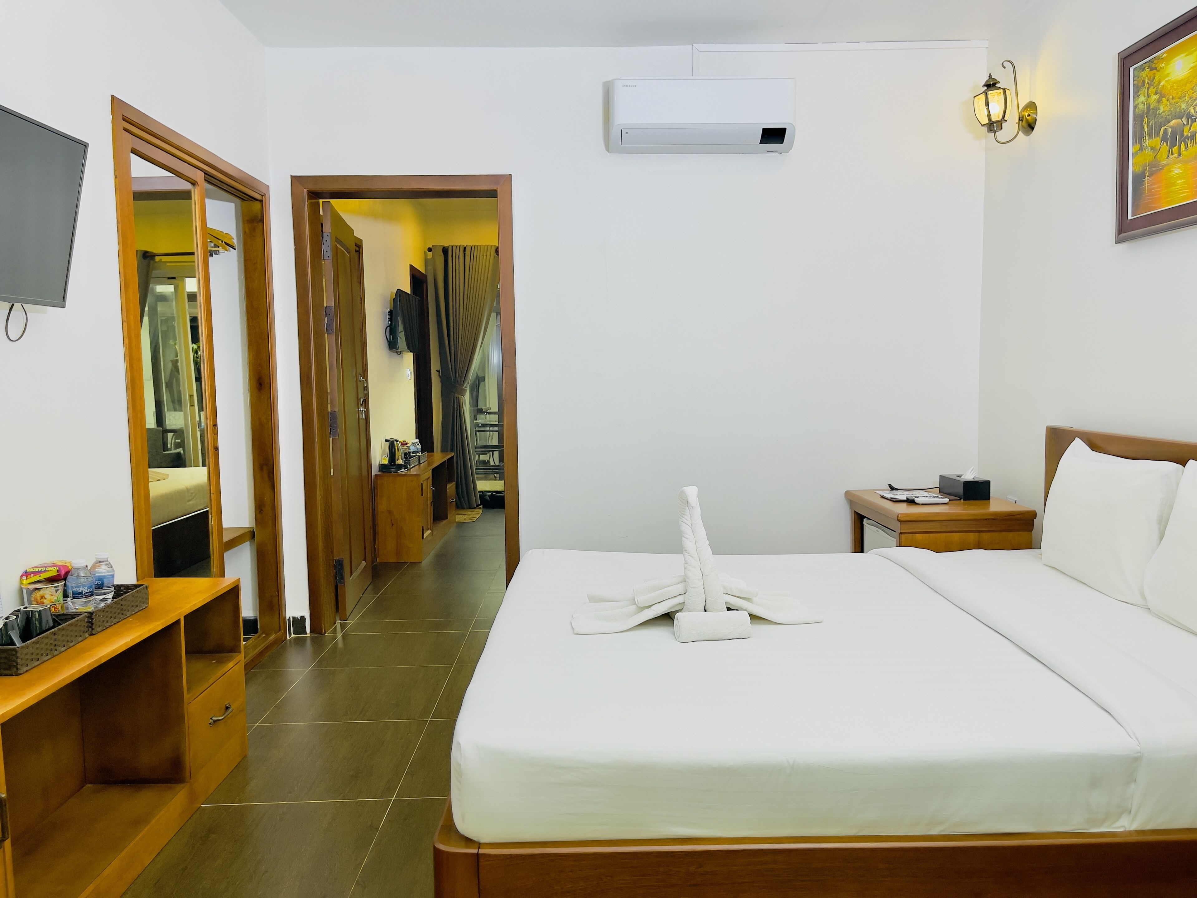 Une chambre d’hôtel équipée d’un lit, d’une table de chevet, d’une télévision, d’un climatiseur et d’un couloir menant à une salle de bain.