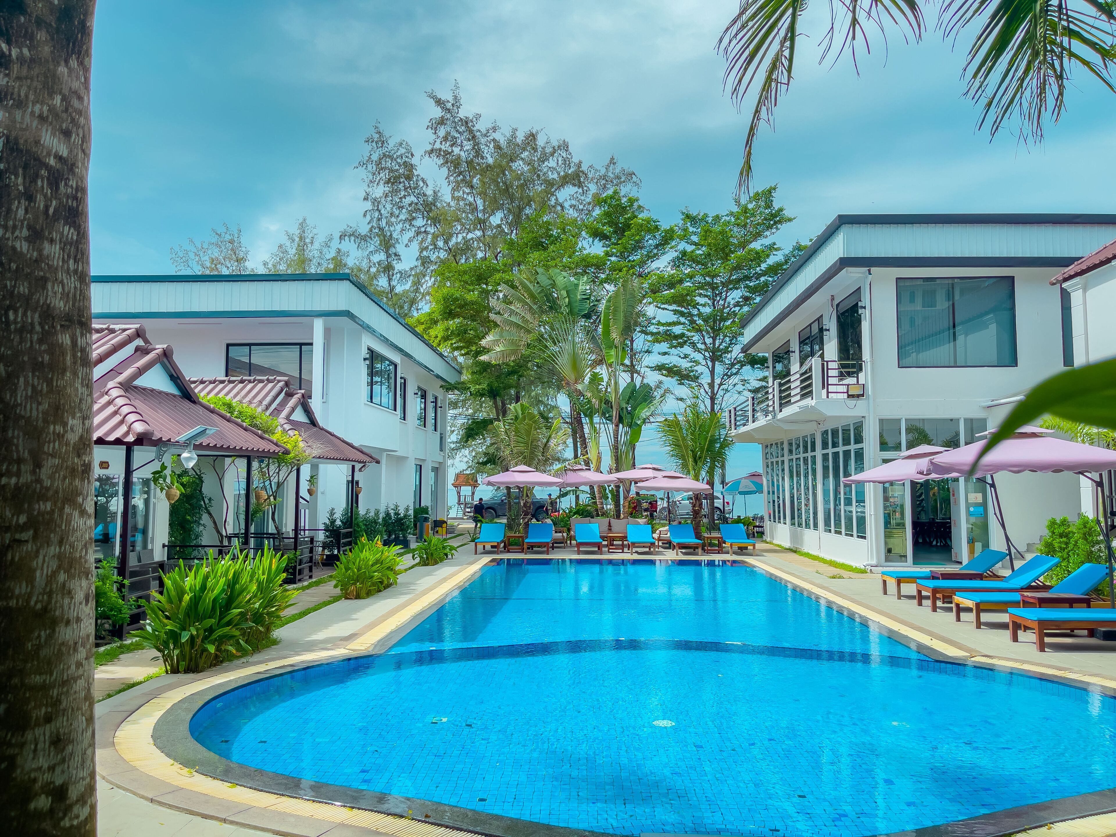 A swimming pool surrounded by lounge chairs and umbrellas, with buildings and trees in the background.