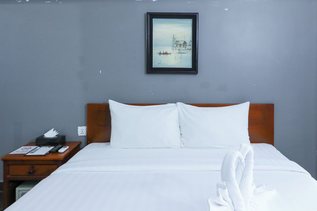 A neatly made bed with white linens and pillows, a wooden headboard, a nightstand with a remote control and a phone, and a framed picture on the wall.