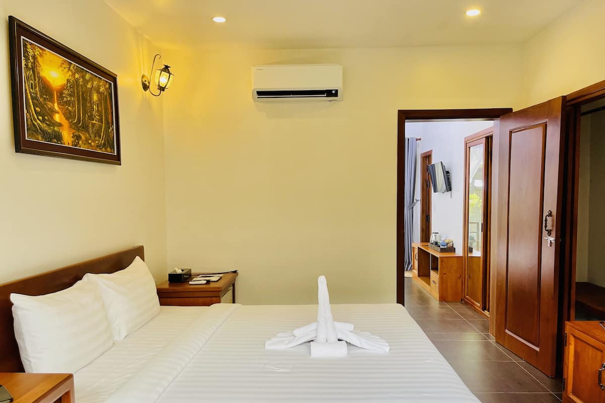 A hotel room with a neatly made bed, a bedside table with a lamp, an air conditioning unit, and a painting on the wall.