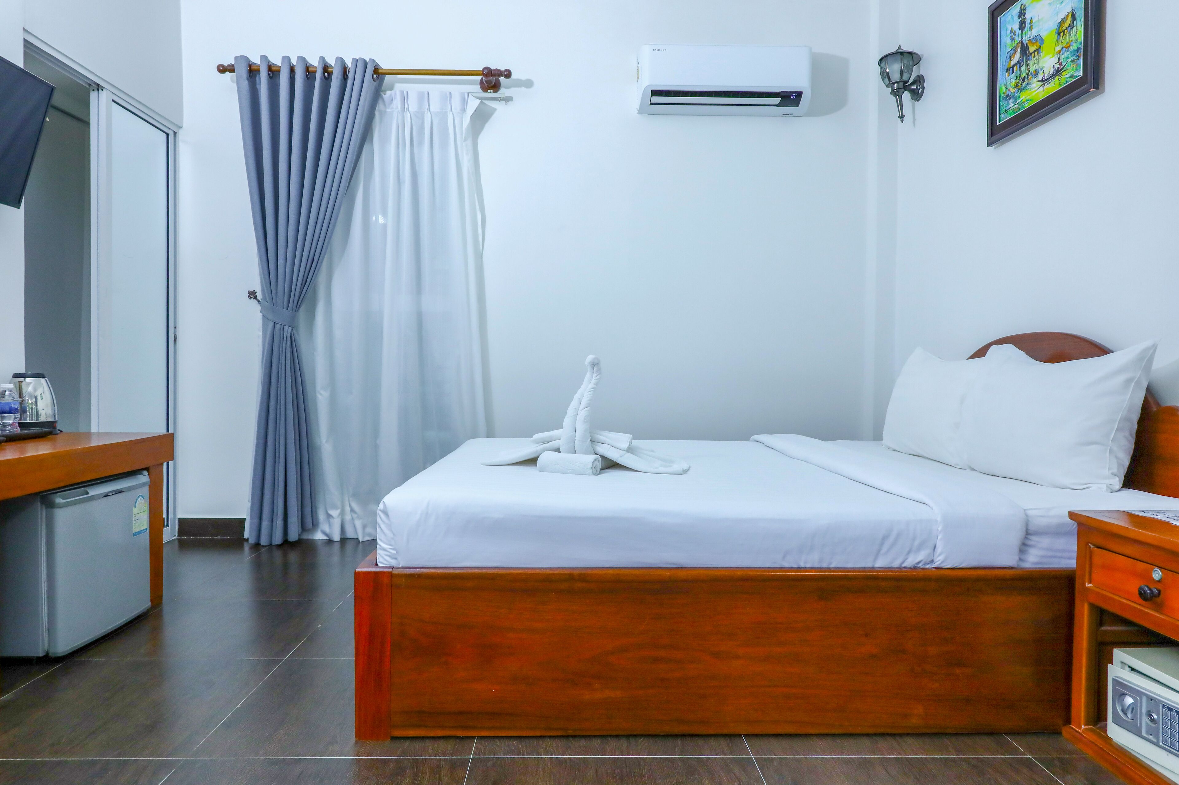 A hotel room with a wooden bed, white bedding, a desk with a computer, an air conditioner, and a wall-mounted TV.