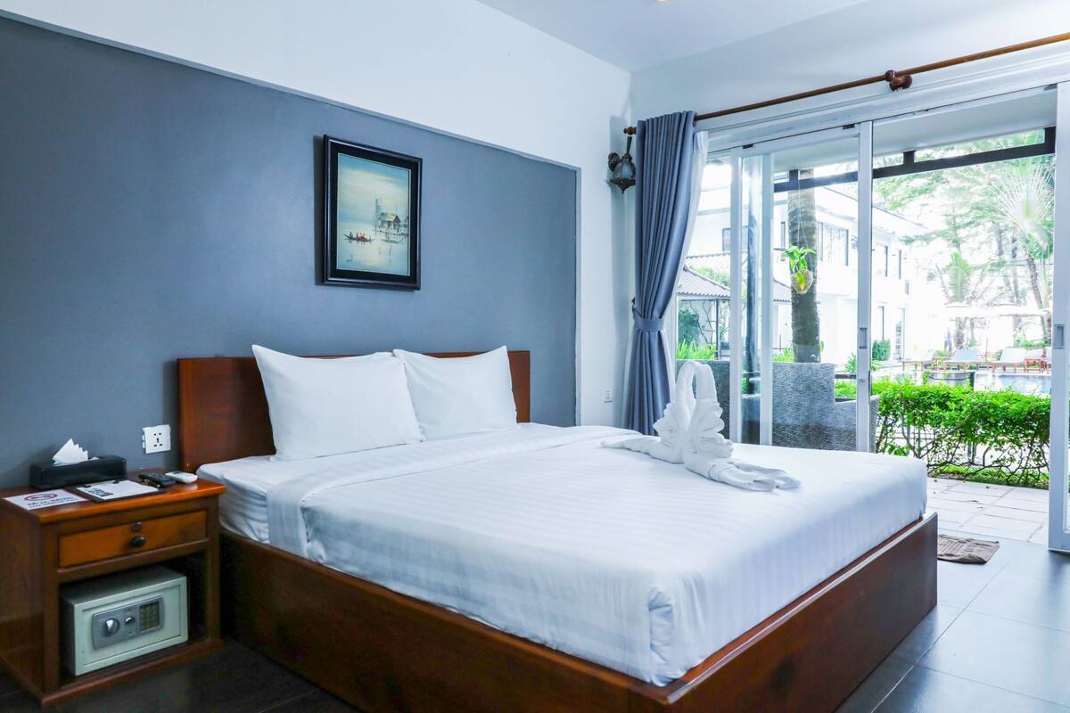 A hotel room with a large bed, a nightstand, a bedside table, a wall-mounted painting, and a view of the outdoor area through a glass door.