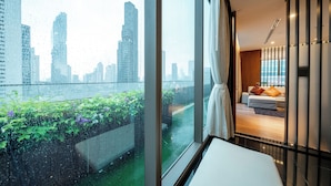 Habitación de hotel moderna con un gran ventanal que da a un paisaje urbano y un balcón con vegetación.