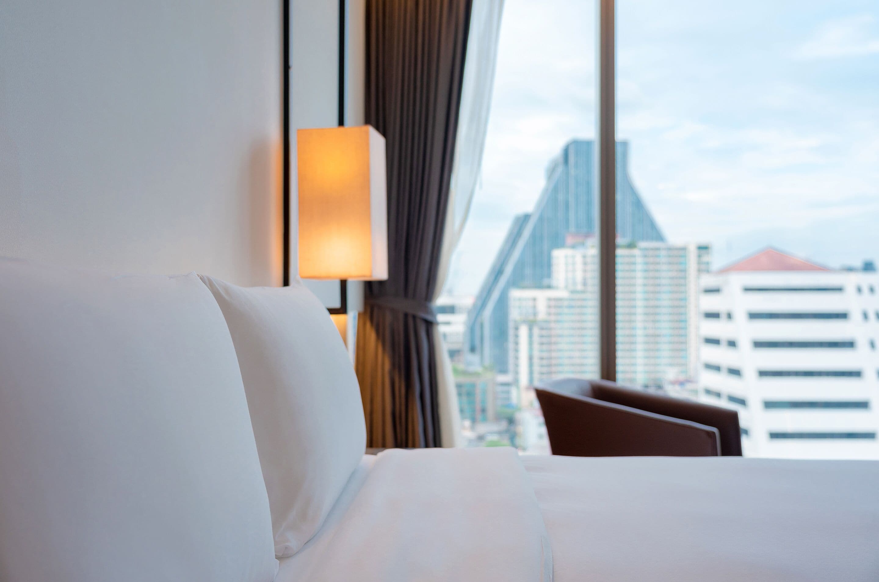 A hotel room with a bed, a lamp, and a view of a cityscape through large windows.