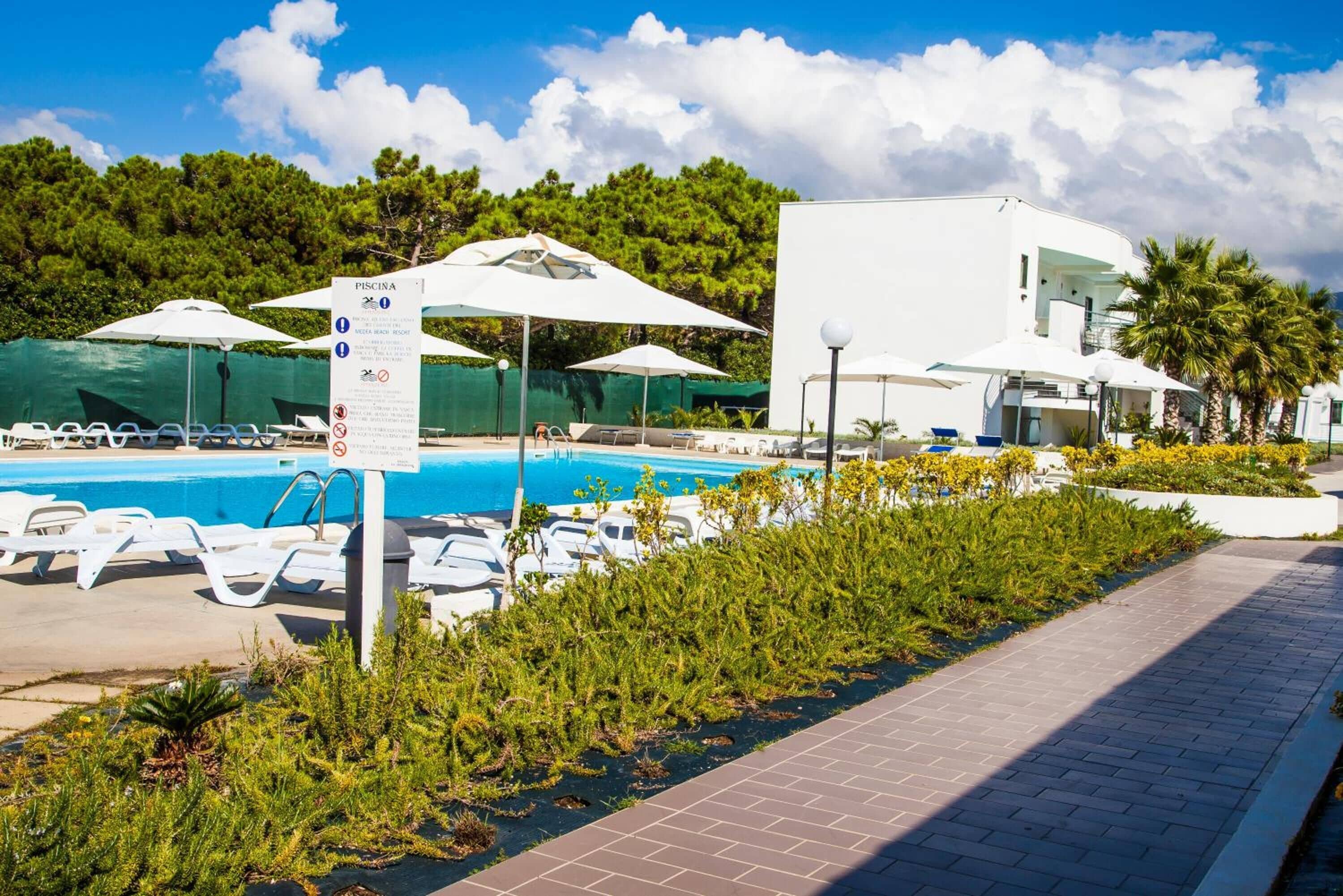 Una piscina con sillas de descanso y sombrillas, rodeada de follaje y un edificio moderno.