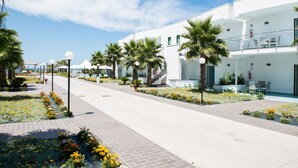 A modern building with a white facade, a covered outdoor area with seating, and a pathway lined with palm trees and flower beds. - Medea Beach Resort (Capaccio-Paestum)