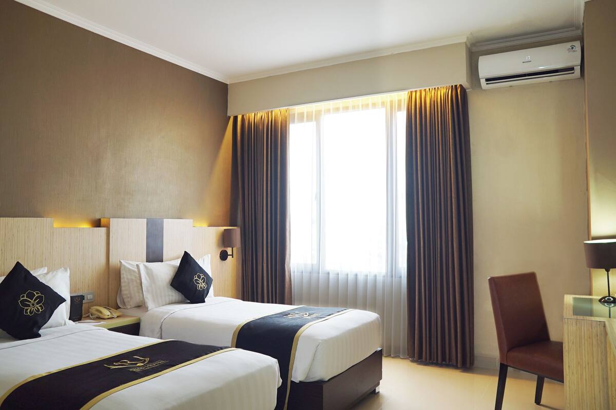 A hotel room with two beds, a desk, a chair, an air conditioning unit, and a window with curtains.
