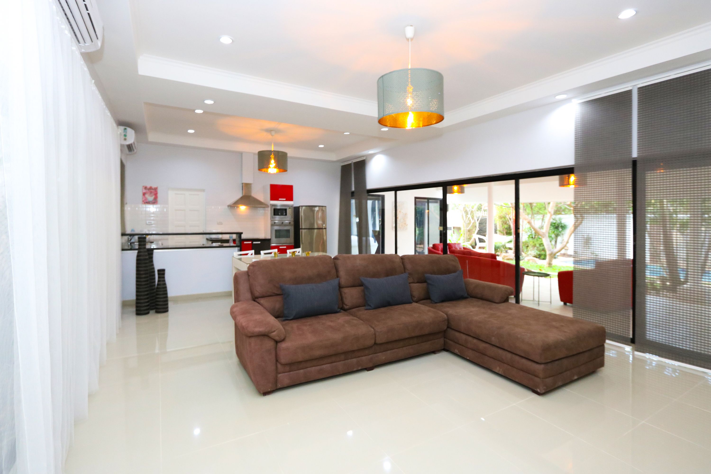 A modern living room with a brown sectional sofa, a kitchen in the background, and large windows offering a view of the outdoors.