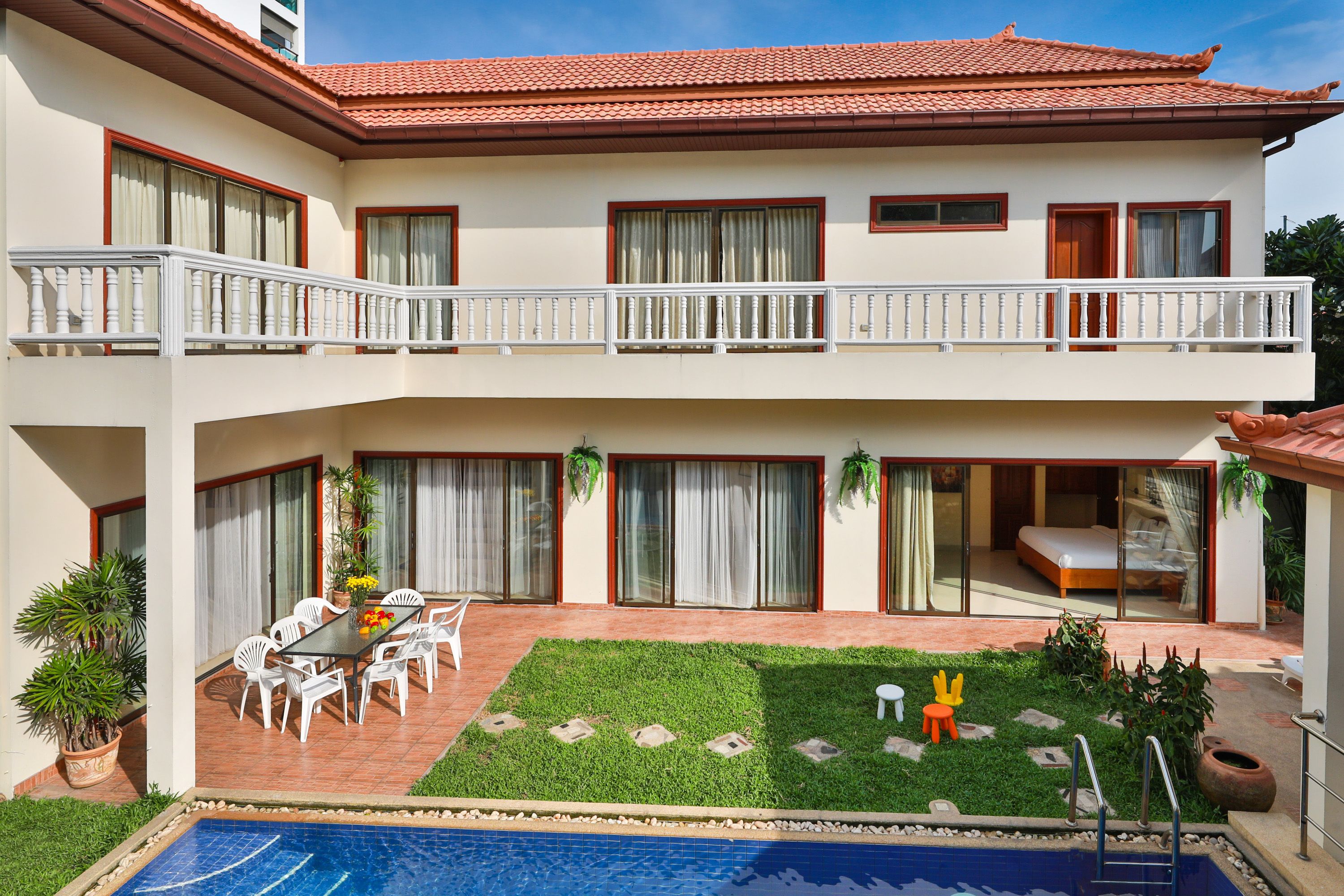 A two-story building with a swimming pool, a dining area with outdoor chairs, and a small garden with a statue.