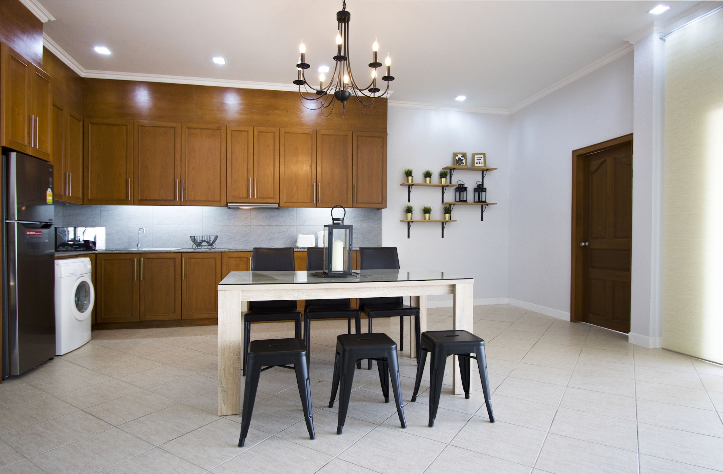 Une cuisine moderne avec des meubles en bois, un réfrigérateur en acier inoxydable, un lave-linge, une table à manger avec quatre tabourets noirs et un lustre.