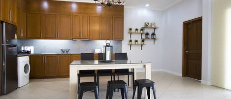 Une cuisine moderne avec des meubles en bois, un réfrigérateur en acier inoxydable, un lave-linge, une table à manger avec quatre tabourets noirs et un lustre.
