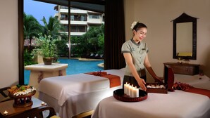 Une salle de spa équipée de deux tables de massage, d’une petite table avec une bougie, et offrant une vue sur une piscine et des palmiers à l’extérieur.