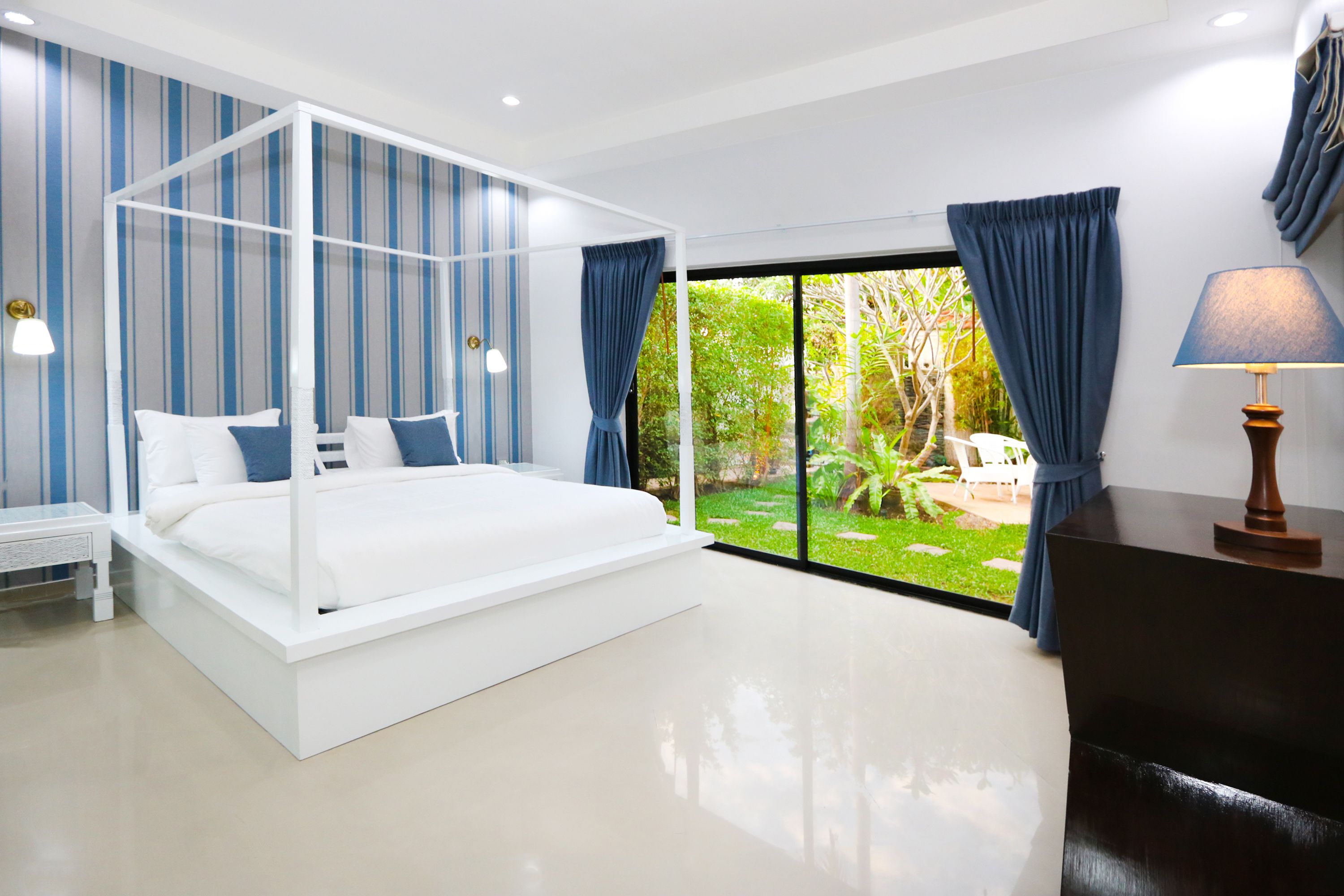 A bedroom with a four-poster bed, a nightstand, a desk with a lamp, and a view of a garden through a large window.