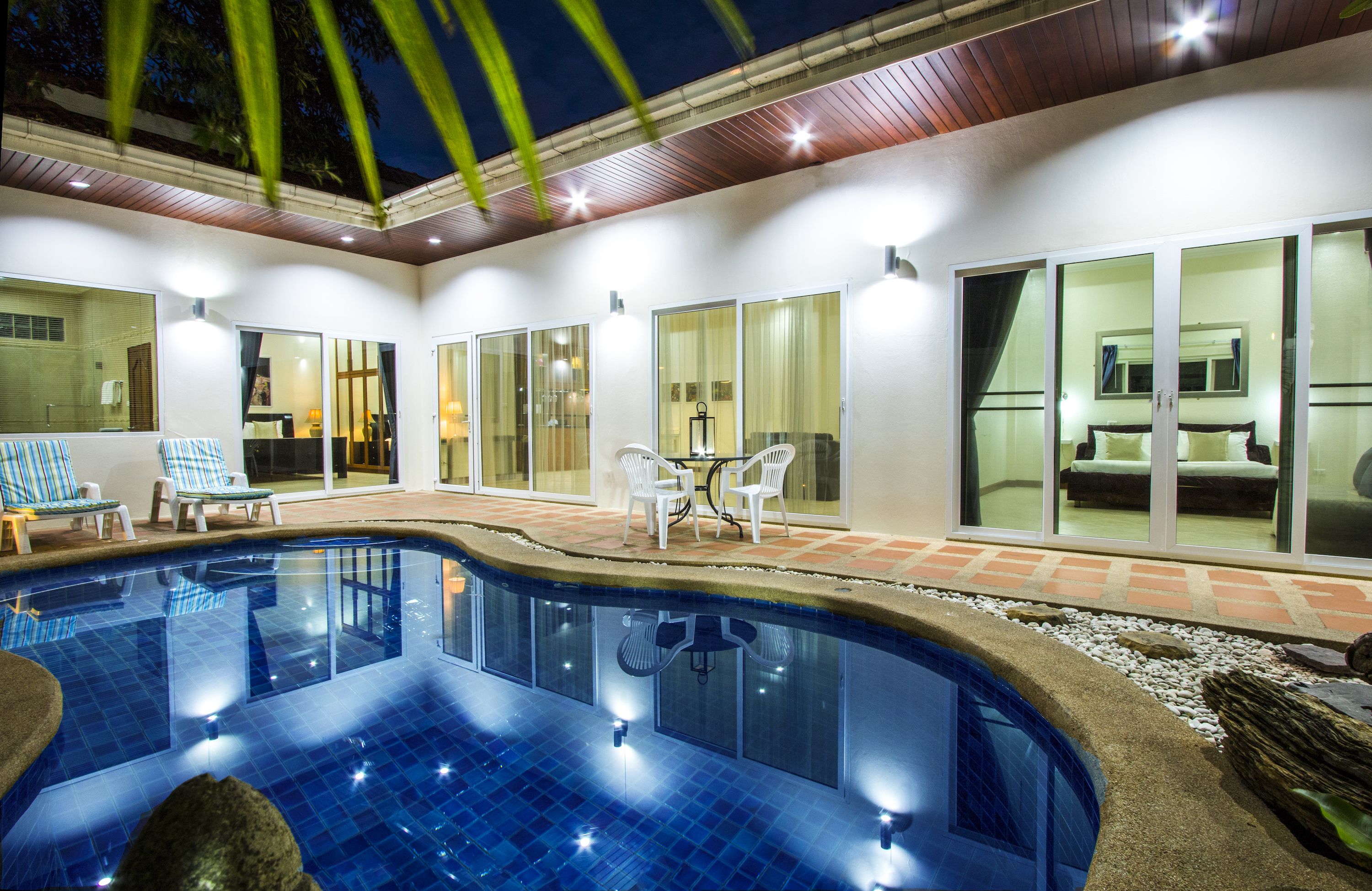 A swimming pool with lounge chairs and a table, surrounded by glass doors and a patio area.